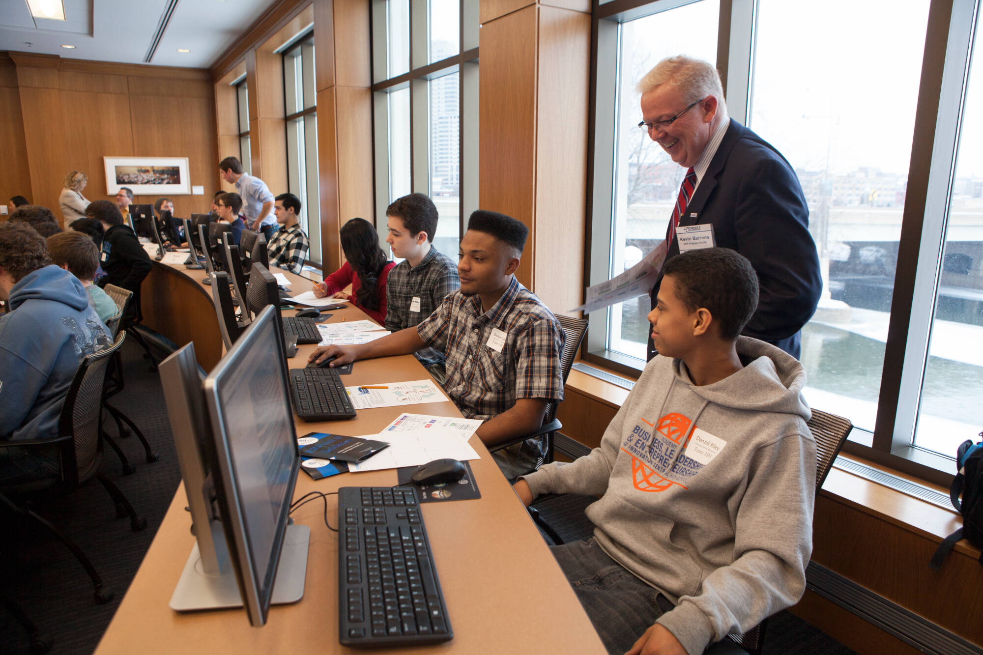 Instructor standing beside students working at desktop computers in a bright classroom with large windows and city view.